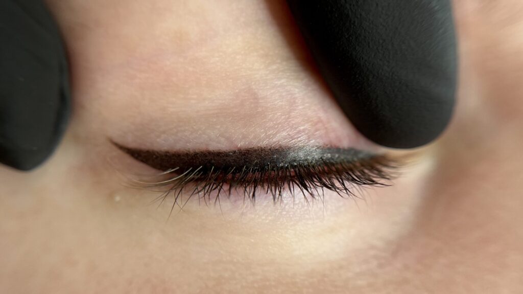 This photo is of a close up image of a classic permanent eyeliner. The eye is closed, and a black glove is pulling the eye taught. The eyeliner is sleek and defined, tapering into a subtle wing to elongate the eye.
