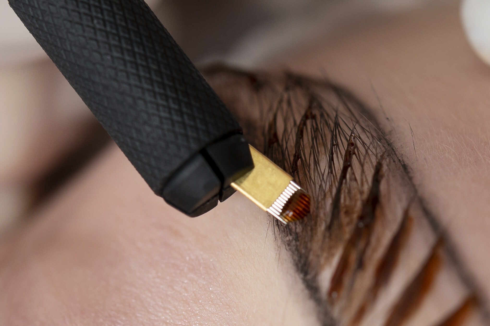 Close-up of a cosmetic tattoo procedure showing a technician using a microblading tool to create fine, hair-like strokes on a client&rsquo;s eyebrow while wearing protective gloves.